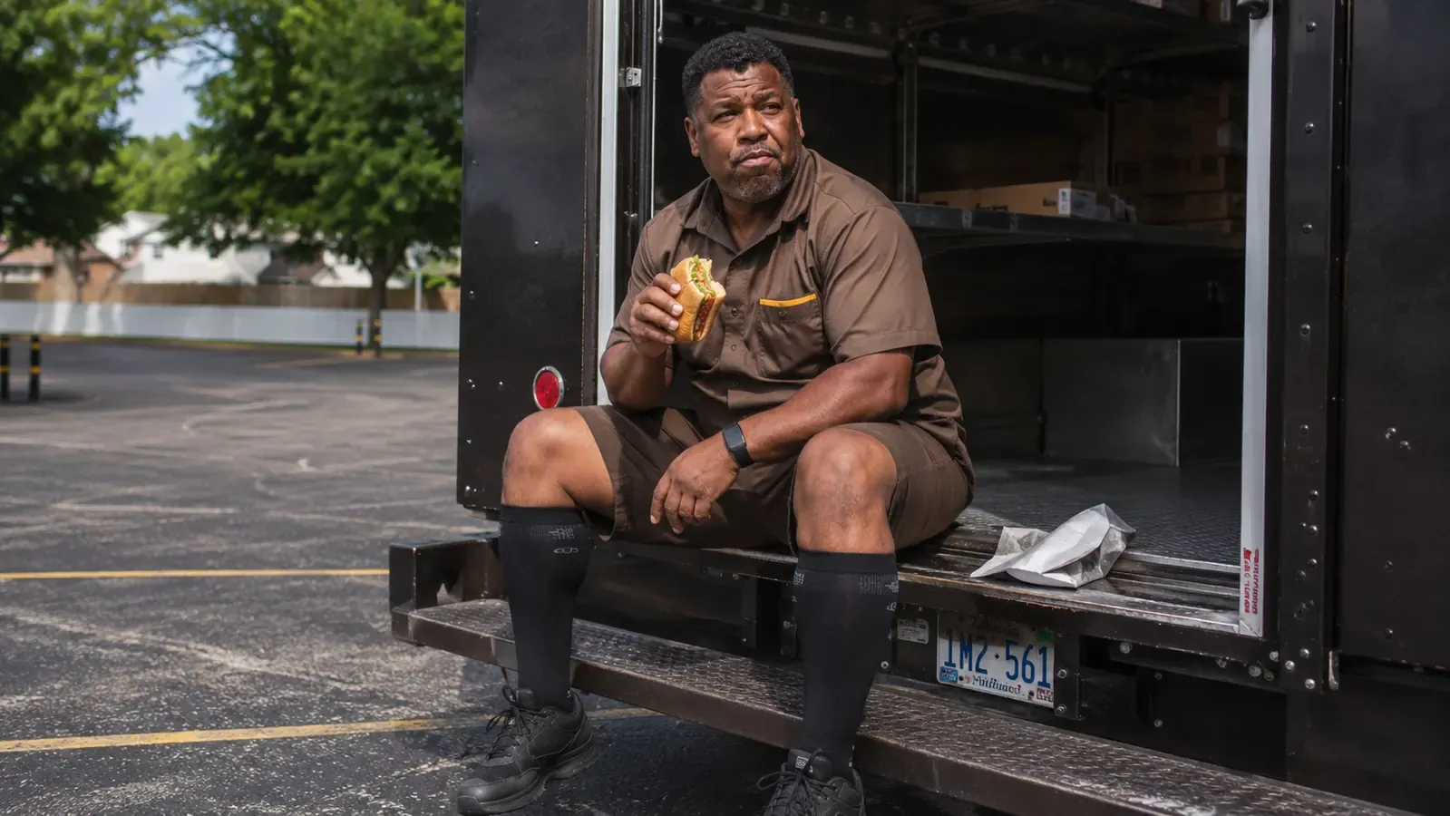 Delivery driver seated on truck step eating lunch, wearing compression socks, relaxed mid-day rest stop scene