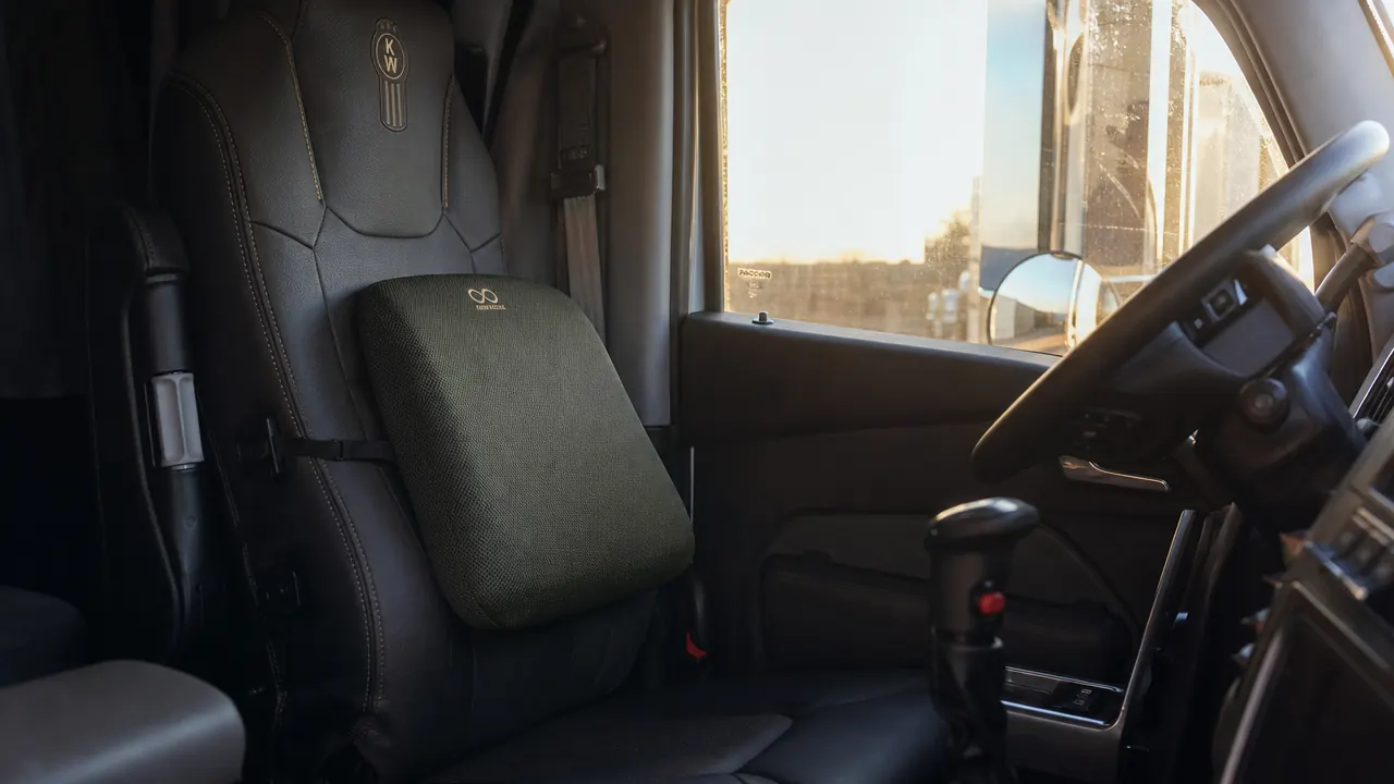 Truck driver's cab seat with a green memory foam lumbar support pillow attached via straps, morning light through the windshield